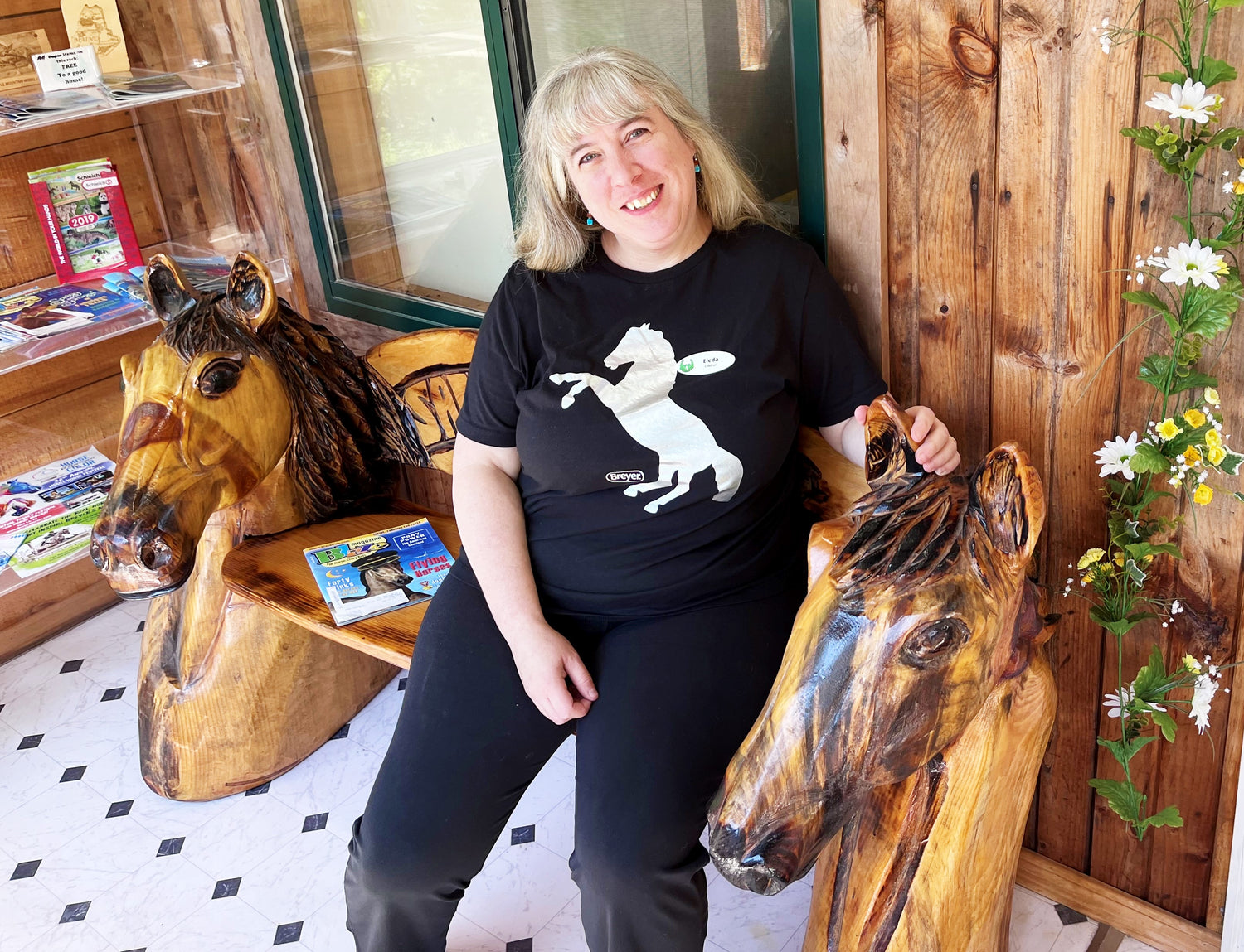 Eleda smiling, sitting on carved wood bench in the store foyer that has a horse head as each end.
